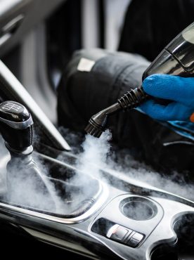 A person is using a steam cleaner inside a car, focusing on the gear shift area, releasing steam to eliminate stains and disinfect surfaces effectively.